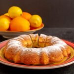 Cytrynowa babka bezglutenowa z cukrem pudrem na talerzu, w tle miska z cytrynami