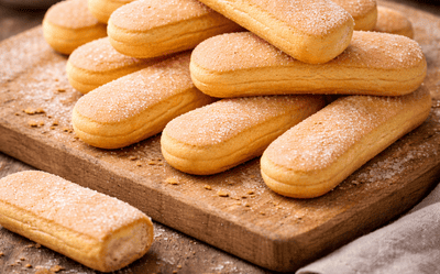 Jasne biszkopty ułożone w stos na drewnianej desce, lekko oprószone cukrem, widoczne okruszki, w tle miseczka z truskawkami i kuchenne akcesoria w domowym otoczeniu.