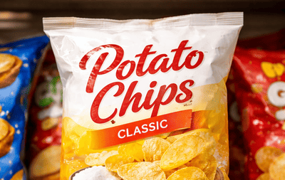 Paczka klasycznych chipsów ziemniaczanych stojąca na sklepowej półce wśród innych przekąsek, lekko nierówne opakowanie, realistyczne oświetlenie.
