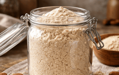 Szklany słoik z jasnobeżową mąką migdałową na kuchennym blacie, wokół rozsypane migdały i mąka, ciepłe naturalne światło i lekko nieuporządkowana domowa sceneria.