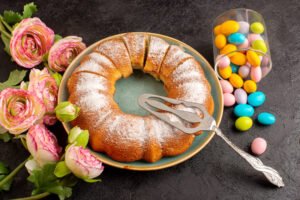 zdjęcie babki wielkanocnej bez glutenu posypanej cukrem pudrem w otoczeniu wiosennych kwiatów i malutkich kolorowych pisanek wielkanocnych