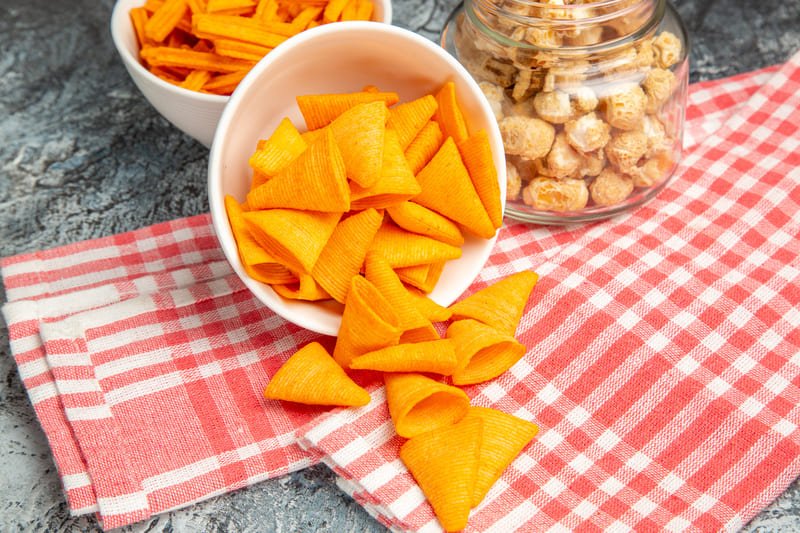 Chrupiące przekąski bez glutenu – chrupki serowe w miseczce oraz popcorn w słoiku na kuchennym blacie.