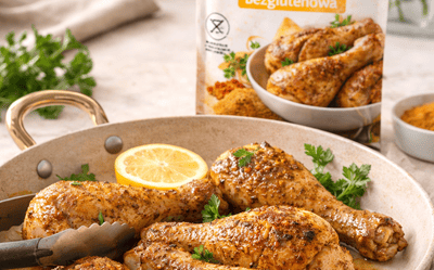 Pieczone pałki z kurczaka z przyprawą bezglutenową w naczyniu na stole, w tle rozmyte opakowanie przyprawy i świeże zioła