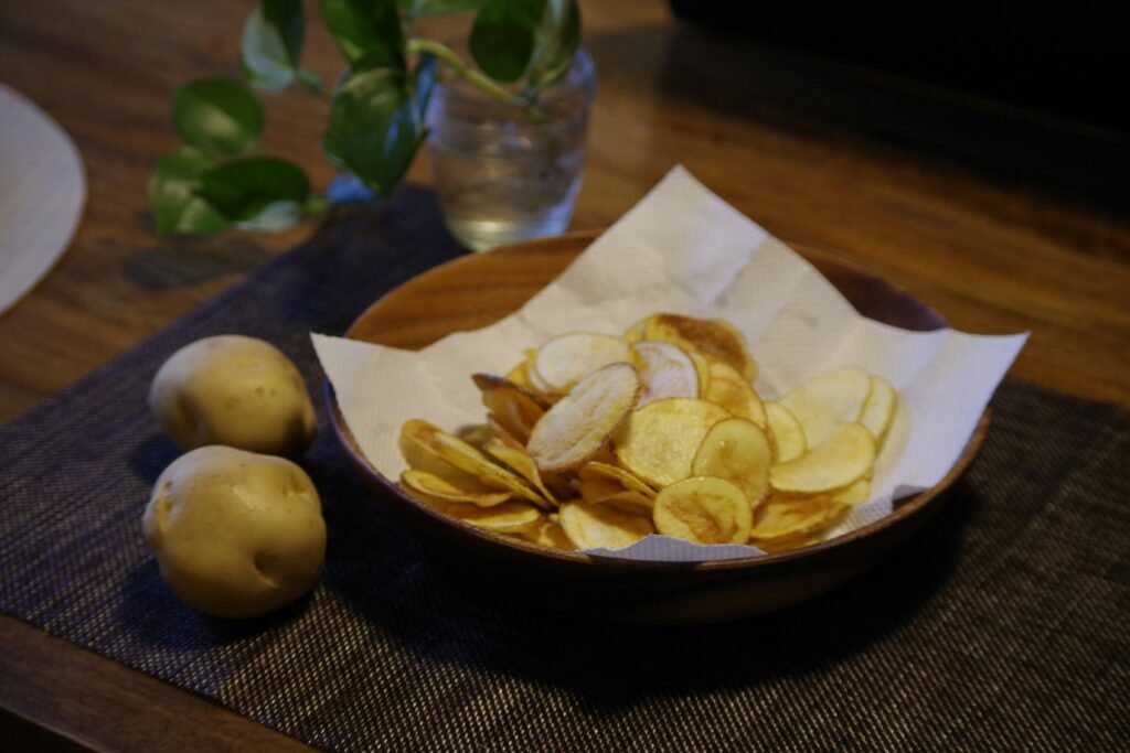 chipsy bezglutenowe djęcie bezglutenowych chipsów w miseczce na ręczniku papierowym, ustawione na stole z dwoma ziemniakami obok miski