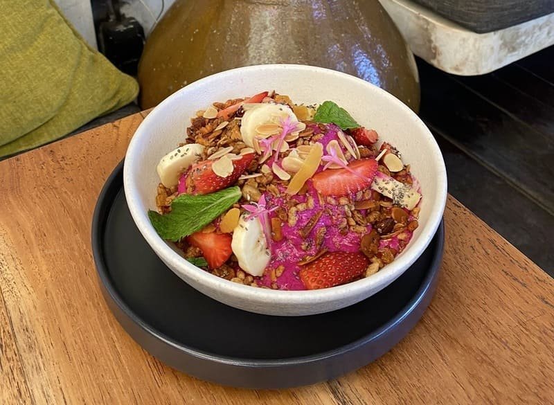 Miseczka smoothie bowl w intensywnym różowym kolorze, udekorowana truskawkami, bananem, granolą bezglutenową, płatkami migdałów, nasionami chia, listkami mięty i jadalnymi kwiatami