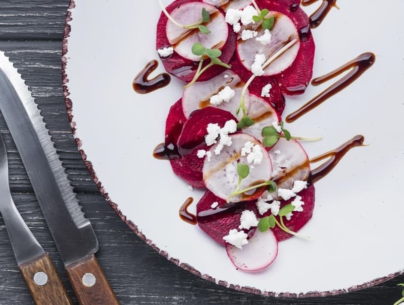 Carpaccio z buraka bez glutenu, podane z rzodkiewką, serem feta, kiełkami i sosem balsamicznym na białym talerzu.