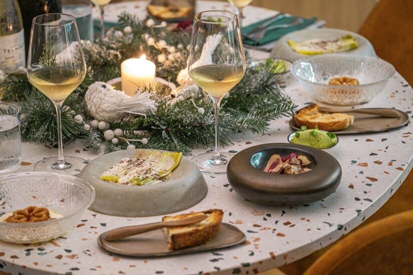 Wigilijny stół z elegancką zastawą, zapalona świeca w stroiku z gałązek świerku, kieliszki z białym winem oraz półmiski z potrawami i pieczywem ustawione na jasnym blacie stołu.