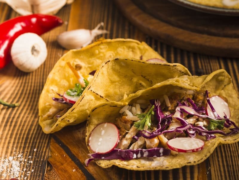 Bezglutenowe tortille kukurydziane wypełnione warzywami, czerwoną kapustą, rzodkiewką i ryżem, podane na drewnianym stole z papryczką chili i czosnkiem w tle.