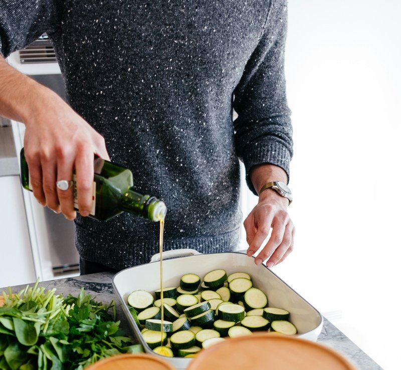 zdjęcie przedstawiające część mężczyzny który polewa oliwą warzywa w naczyniu do zapiekania w kuchni