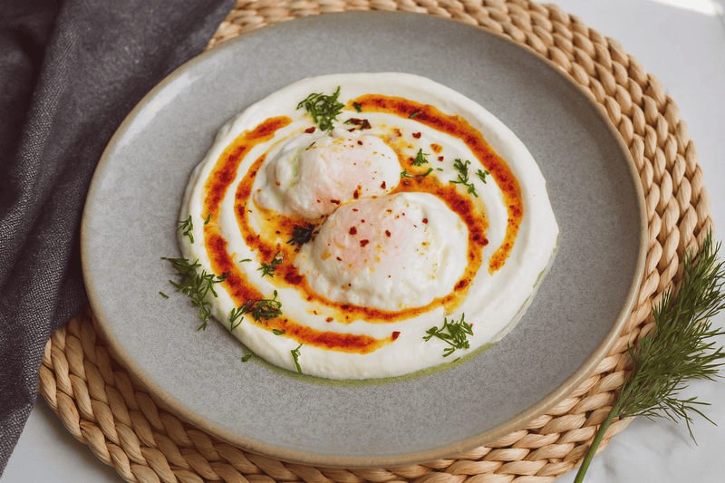 zdjęcie jajek po turecku bez glutenu, na jogurcie, z masłem paprykowym i koperkiem, podane na szarym talerzu, ustawionym na plecionej podkładce na stole