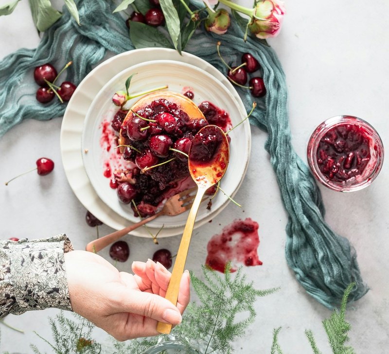 zdjęcie przedstawiające bezglutenowe pancakes z wiśniami i rękę kobiety, która dokłada więcej konfitury