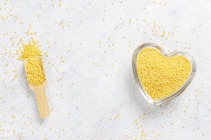 Sucha kasza jaglana w drewnianej misce i łopatce, rozsypana na jasnym, drewnianym blacie.