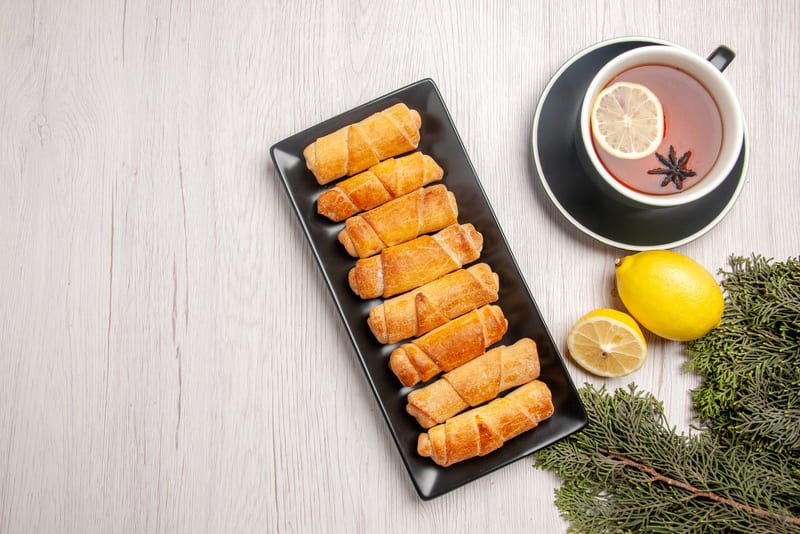 widok-z-gory-ciasto-rurkowe-ciasto-rurkowe-na-czarnym-talerzu-obok-filizanki-herbaty-z-galazkami-cytryny-i-swierku-na-bialym-stole Bezglutenowe paszteciki z kapustą i grzybami ułożone na podłużnym talerzu na stole obok filiżanki herbaty