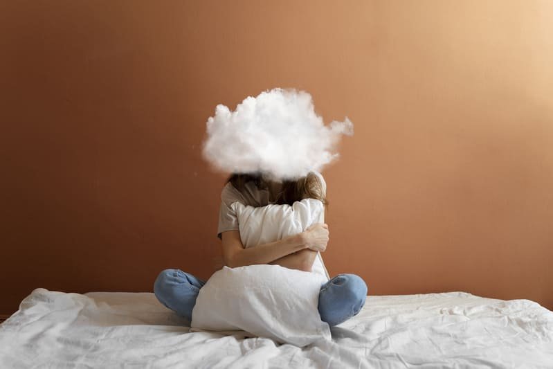 Zatrucie glutenem (1) Osoba siedząca na łóżku, obejmująca poduszkę, z symboliczną chmurą nad głową – ilustracja objawów zatrucia glutenem, takich jak złe samopoczucie, zmęczenie i reakcja organizmu po spożyciu glutenu.