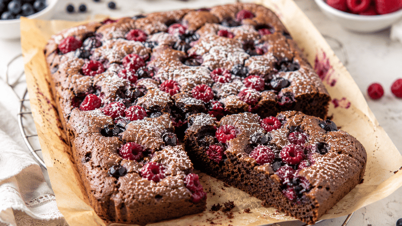 Wilgotne i puszyste ciasto czekoladowe bez glutenu z malinami i borówkami, pieczone z owocami na wierzchu, podane w naturalnym świetle na papierze do pieczenia.